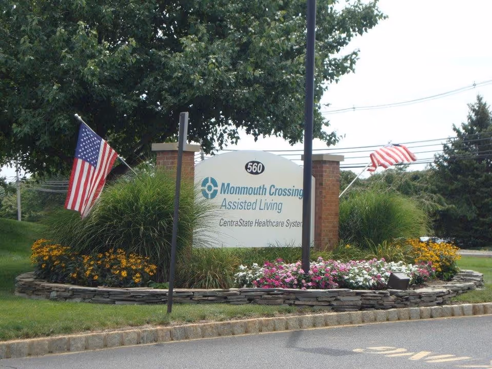 Outdoor view of the entrance sign for Monmouth Crossing Assisted Living, part of CentraState Healthcare System, surrounded by landscaping with flowers, grass, and two American flags on either side.