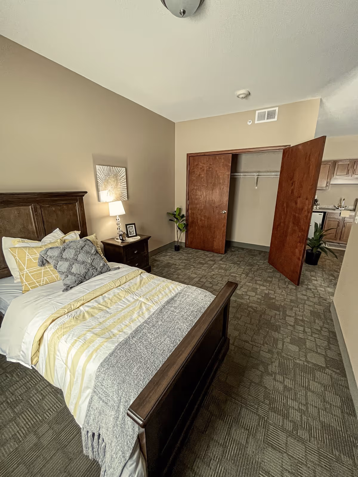A neatly made single bed with yellow and gray bedding in a bedroom. Next to the bed is a wooden nightstand with a lamp, a small framed picture, and a decorative item. The room has beige walls and carpeted flooring. There is an open closet with wooden sliding doors and a potted plant near the closet. In the background, part of a kitchen area with wooden cabinets is visible.