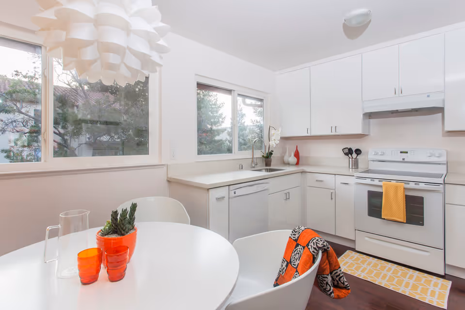 Bright and modern kitchen with white cabinets, a white stove with a yellow towel, a white dishwasher, and a white round dining table with two white chairs. On the table are two small orange glass cups, a clear glass pitcher, and a small potted plant. A colorful orange and black patterned blanket is draped over one chair. Large windows let in natural light, and a unique white pendant light hangs above the table.