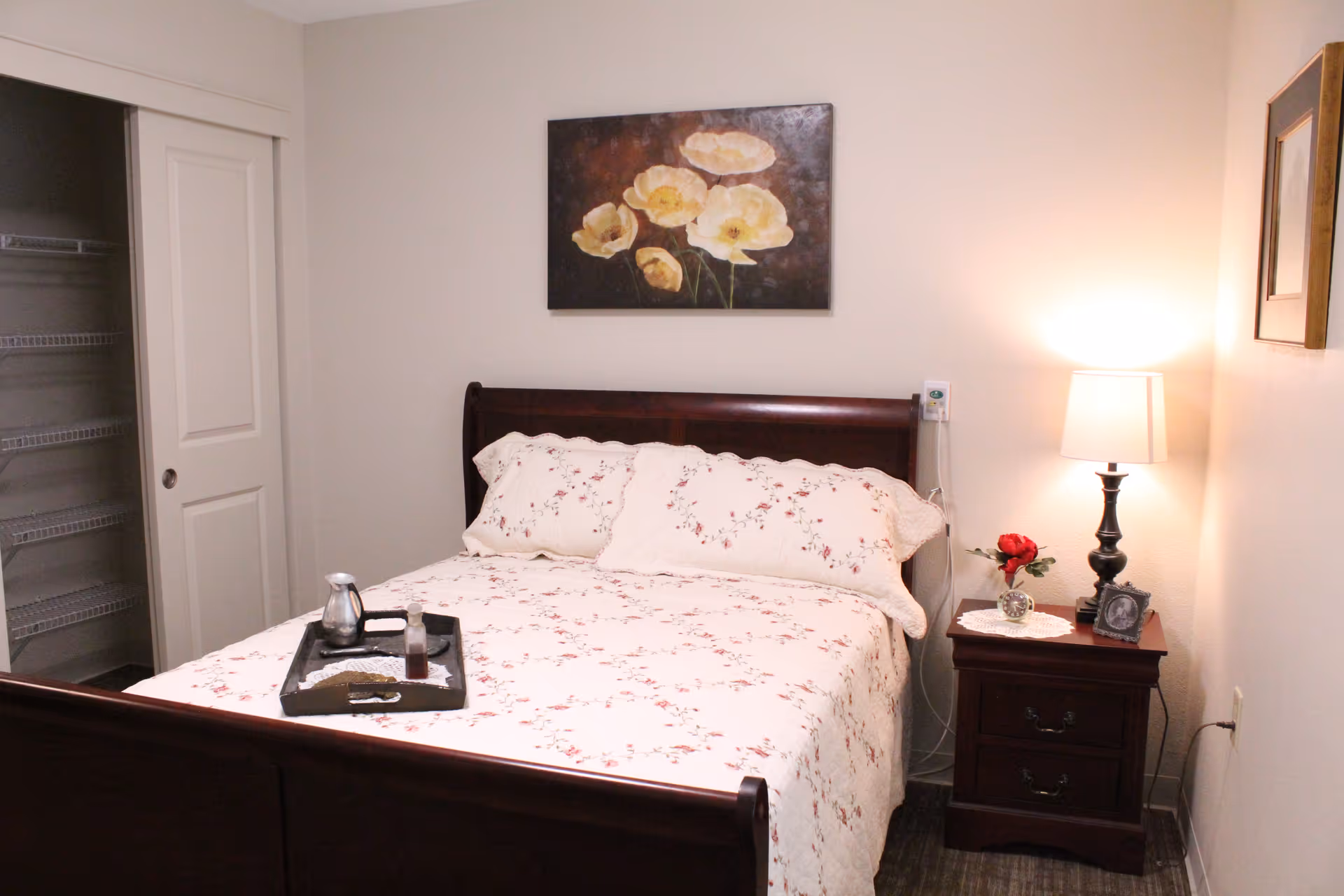 A bedroom with a wooden bed frame and floral bedding. There is a tray with a coffee pot and cup on the bed. A nightstand with a lamp, a small clock, a framed photo, and a red flower in a vase is next to the bed. A floral painting hangs on the wall above the bed, and there is an open closet with wire shelving on the left side.