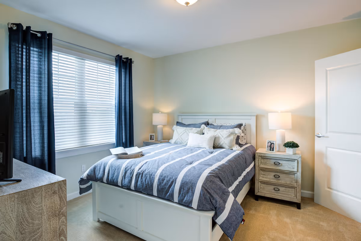 A cozy bedroom with a white bed frame and a blue and white striped comforter. There are multiple pillows on the bed, two bedside tables with lamps, a small plant, and framed photos. A window with dark blue curtains and white blinds is on the left side, and a wooden dresser with a TV is partially visible in the foreground.