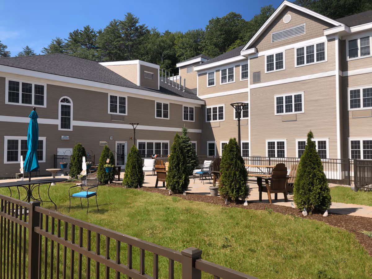 Outdoor patio area at The Prospect-Woodward Health Center at Hillside Village featuring seating arrangements with chairs and tables, small evergreen trees, a closed blue umbrella, and a multi-story beige building with white trim in the background under a clear blue sky.