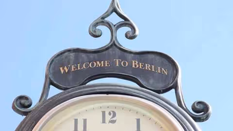 Decorative arched clock above a 'Welcome to Berlin' sign against a blue sky.