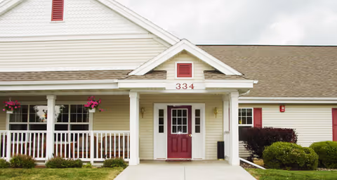Front entrance of a single-story assisted living building with a covered porch, white columns, a red door and the number '334' above it.