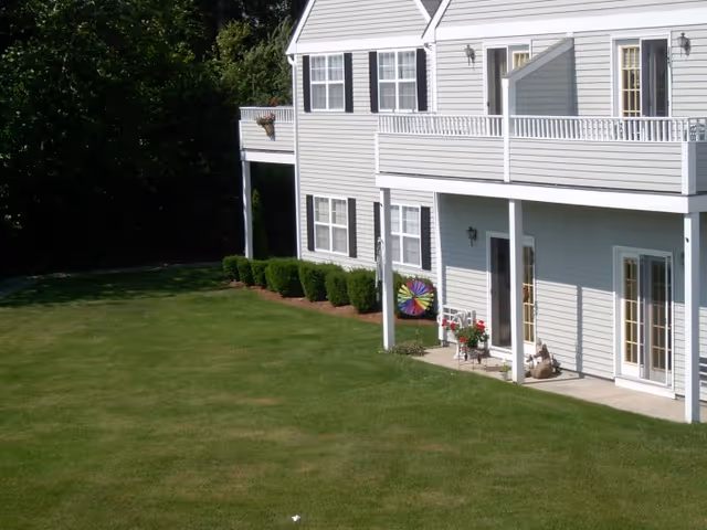 Exterior view of a light gray two-story building with white trim, featuring a balcony and patio area. The building is surrounded by a well-maintained green lawn and some bushes along the side. There is a colorful pinwheel and some potted plants on the patio.
