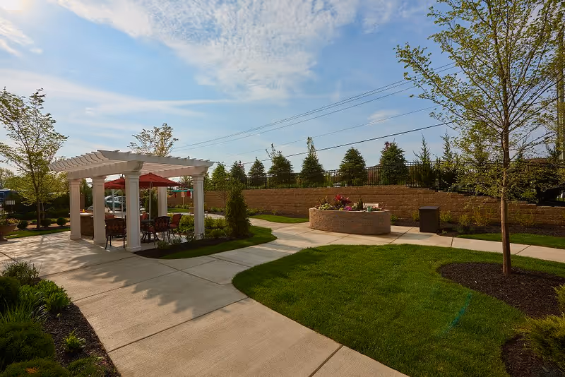 Outdoor area of Dublin Retirement Village featuring a white pergola with red umbrellas and chairs underneath, surrounded by green grass, trees, and landscaped garden beds, with a clear blue sky overhead.