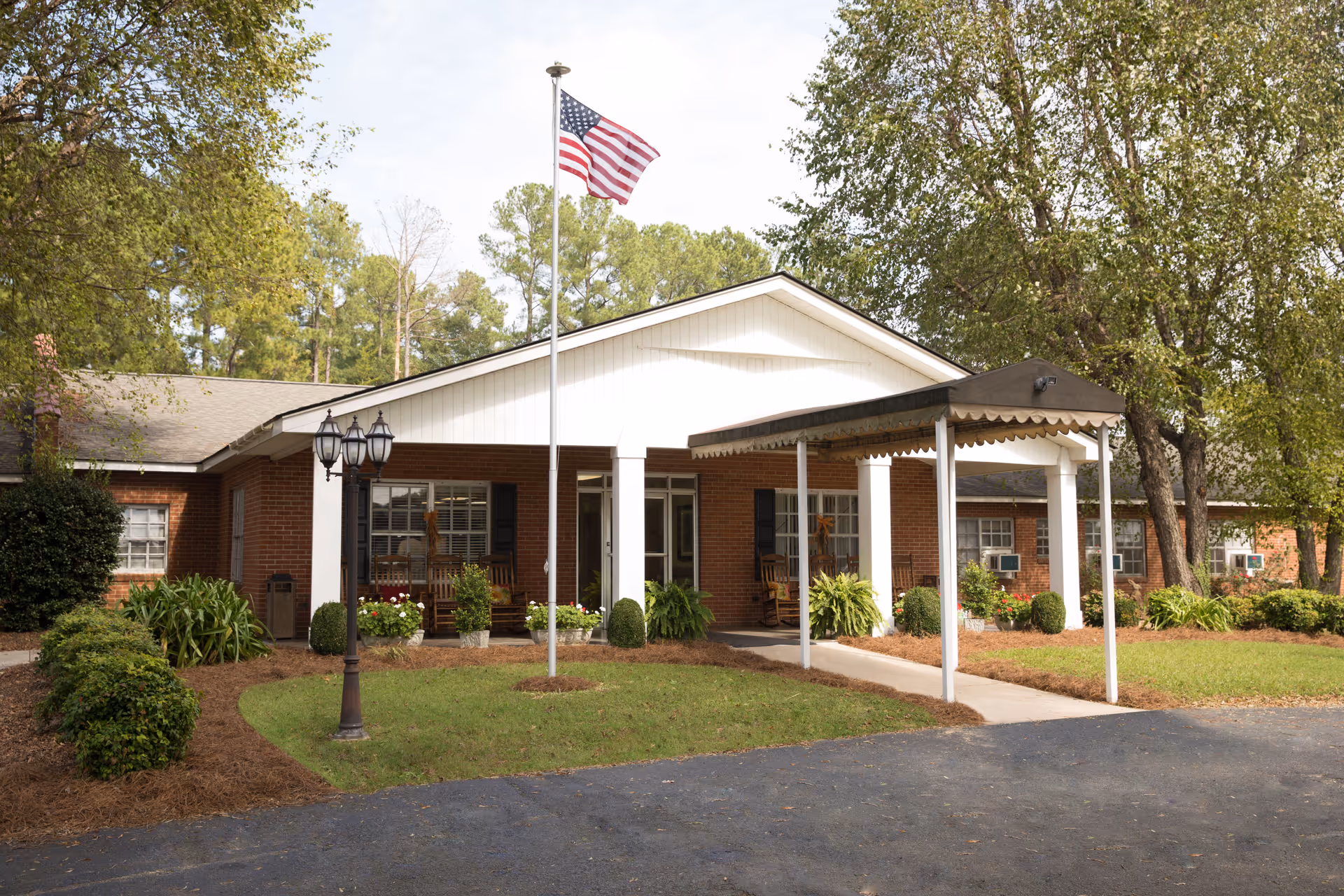 Front exterior view of a single-story brick building with a covered entrance, an American flag on a flagpole, and landscaped greenery including bushes and trees.