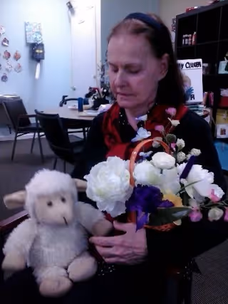 An elderly woman sitting indoors holding a plush sheep toy and a basket of white and purple flowers. Behind her are tables and chairs, a door, and shelves with various items.