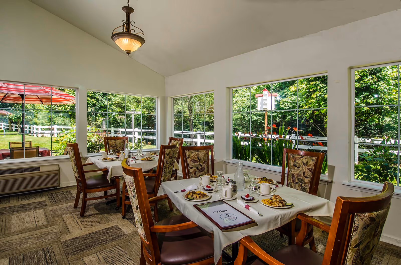 A bright dining room with large windows showing a green outdoor garden. The room has two tables set with white tablecloths, plates of food, cups, and a menu. The chairs have wooden frames with patterned upholstery. A hanging light fixture is visible on the ceiling.