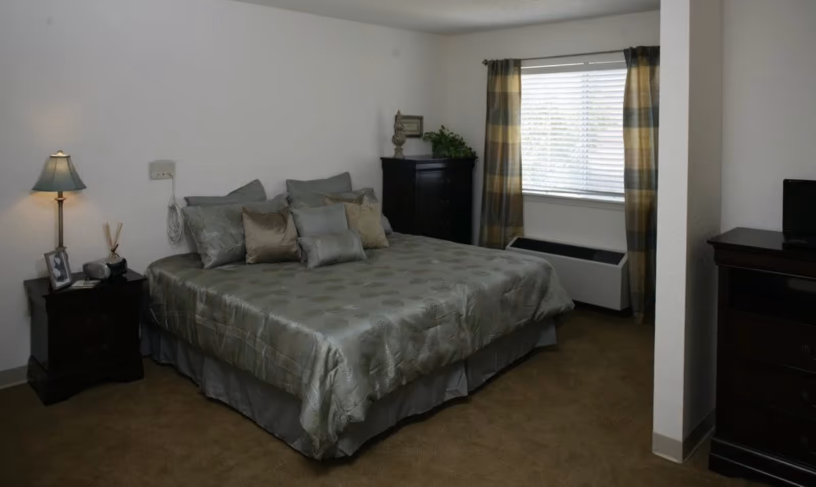 Neatly furnished bedroom with a large bed, nightstands, dresser, lamp, and a window with curtains.