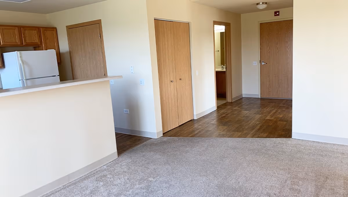 Interior view of an empty apartment unit at Poplar Creek Village showing a kitchen area with wooden cabinets and a white refrigerator, a carpeted living space, and a hallway with wooden doors leading to other rooms.