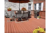 Sunlit wooden deck patio with a table, chairs and umbrella, potted plants, and a gas grill beside a brick and siding building.