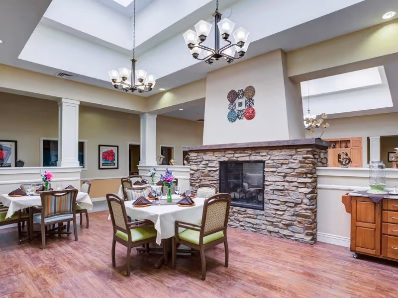 A dining room in a senior living facility with round tables covered in white tablecloths, each set with napkins, glasses, and floral centerpieces. The room features a stone fireplace in the center, wooden flooring, and two chandeliers hanging from a ceiling with skylights. There are framed artworks on the walls and a wooden cabinet with a water dispenser on the right side.