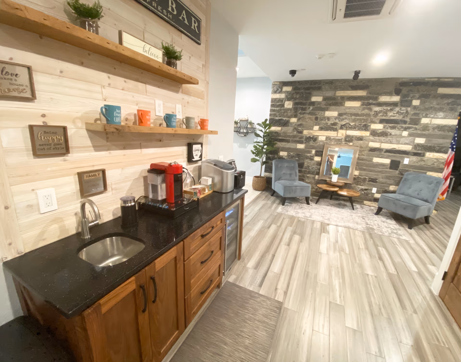 Interior view of a cozy common area in a senior living facility featuring a small kitchenette with a sink, coffee maker, and shelves holding colorful mugs. Adjacent to the kitchenette is a seating area with two blue upholstered chairs, a small wooden coffee table, a decorative mirror, a potted plant, and a stone accent wall. The floor is covered with light wood-style tiles.