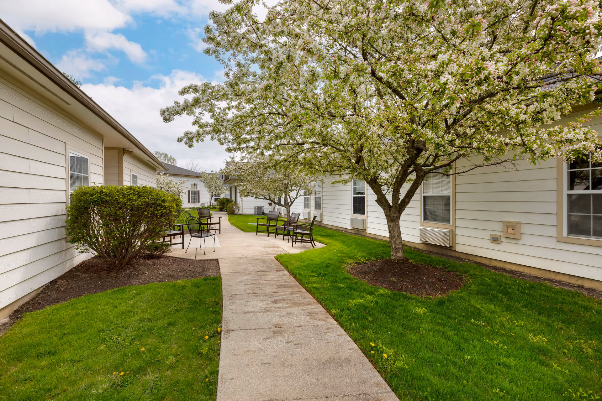 Outdoor walkway between buildings at Lorain Estates Senior Living with green grass, blooming trees, and several black metal chairs and tables along the path under a partly cloudy sky.