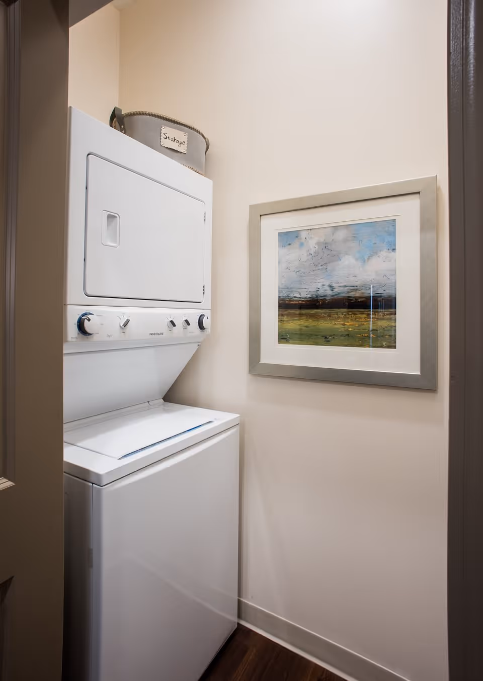 A small laundry area with a stacked white washer and dryer unit. On top of the dryer is a gray fabric storage basket labeled 'Storage'. A framed landscape painting with a silver frame hangs on the adjacent wall. The walls are painted light beige and the floor is dark wood.