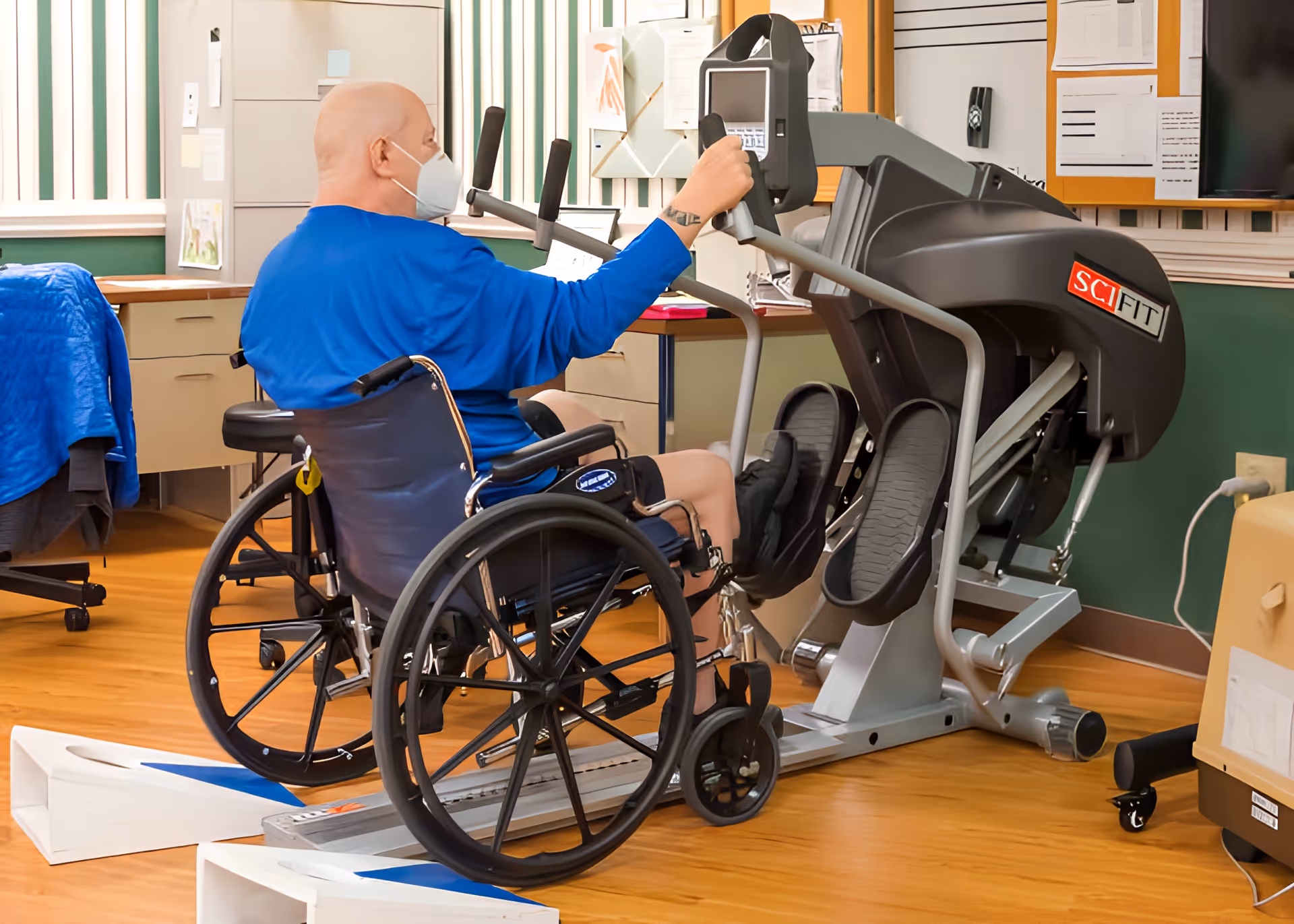 A person in a wheelchair wearing a blue shirt and a face mask is using a SCI FIT exercise machine in a room with wooden floors, desks, and office chairs.