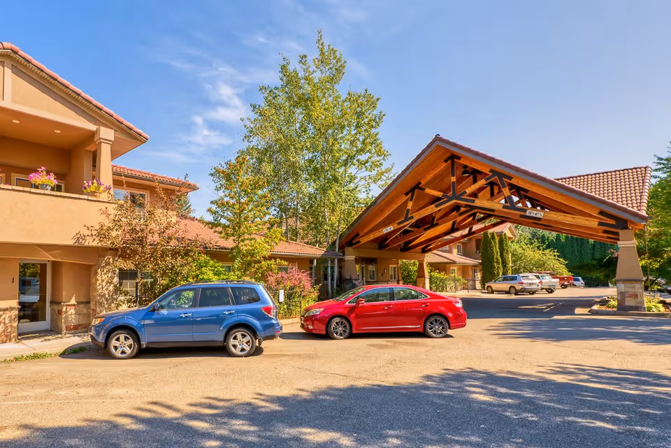 Exterior view of Courtyard at Coeur d'Alene senior living facility showing a covered entrance with wooden beams, parked cars, and a building with balconies and flower pots under a clear blue sky.