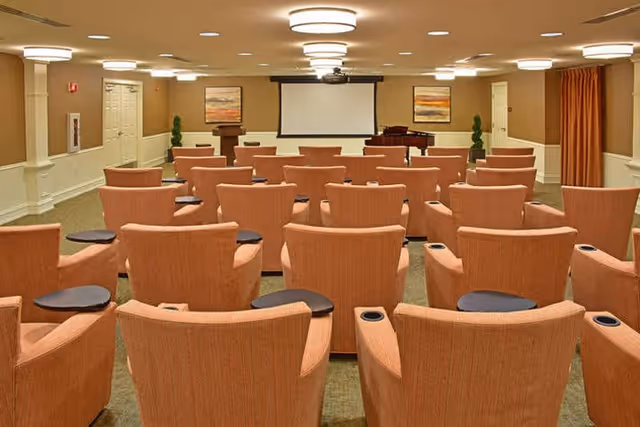A conference or presentation room with multiple rows of orange upholstered chairs, each equipped with a small black side table and cup holder. At the front of the room is a podium, a grand piano, and a large projection screen mounted on the wall. The room is well-lit with ceiling lights and decorated with framed artwork and potted plants.