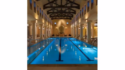 Indoor lap pool in an elegant high-ceilinged room with columns, exposed wooden beams, and seating at the far end.