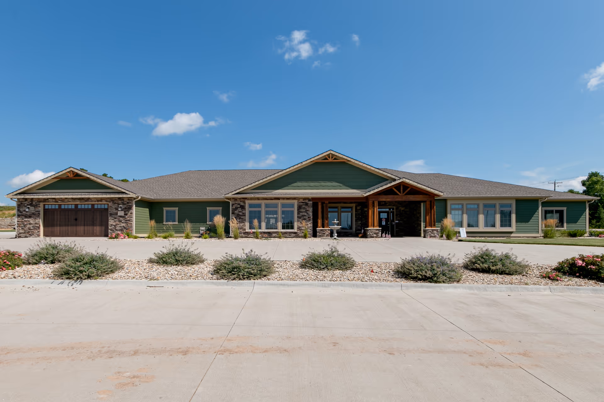 Front exterior view of a single-story building with green siding, stone accents, and a covered entrance with wooden beams. There are multiple windows and a garage door on the left side. The building is surrounded by a concrete driveway and landscaped with bushes and flowers under a clear blue sky.