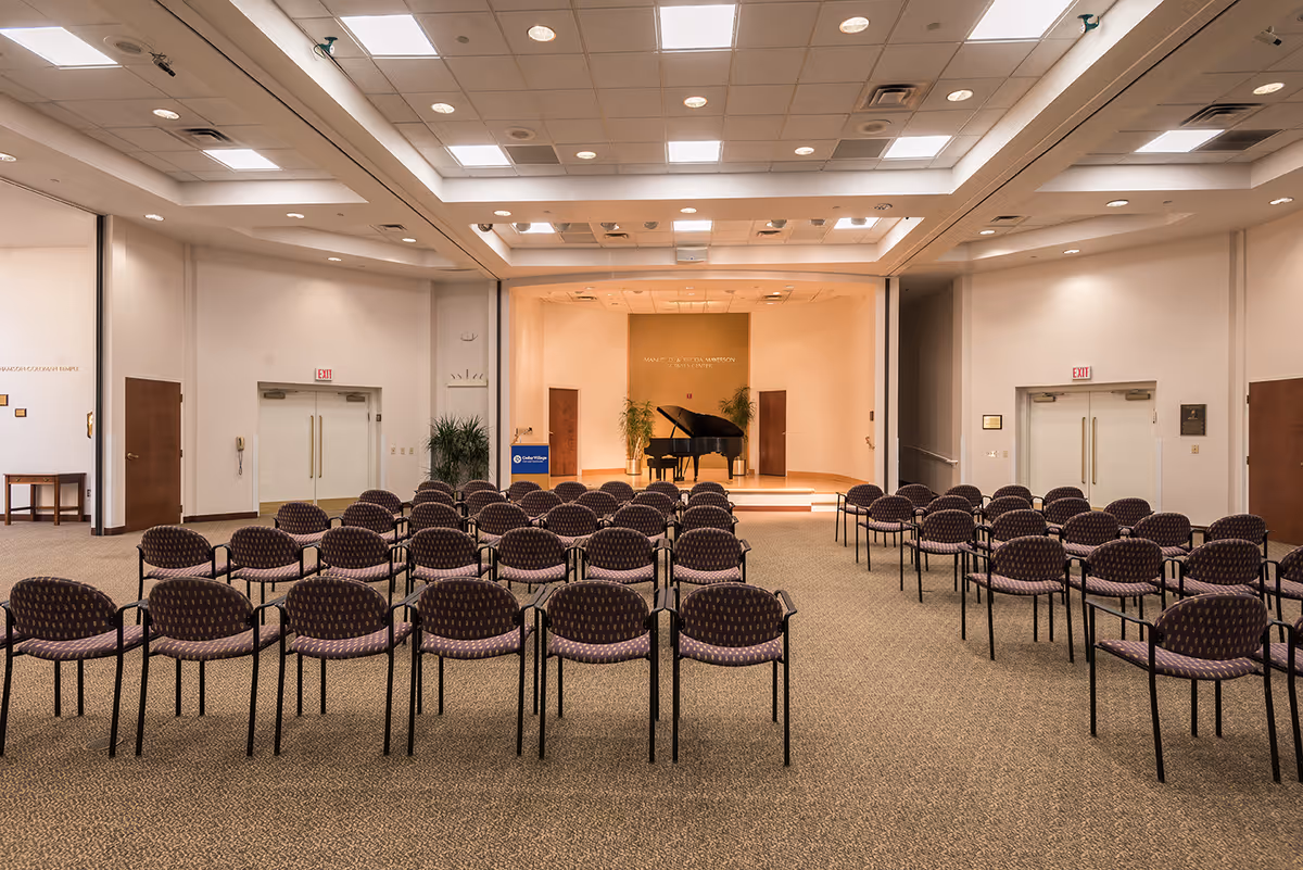 A large room with rows of empty chairs facing a small stage that has a grand piano and two potted plants. The room has a beige carpet, white walls, and a ceiling with recessed lighting and skylights. There are double doors on both sides of the stage and exit signs above the doors.