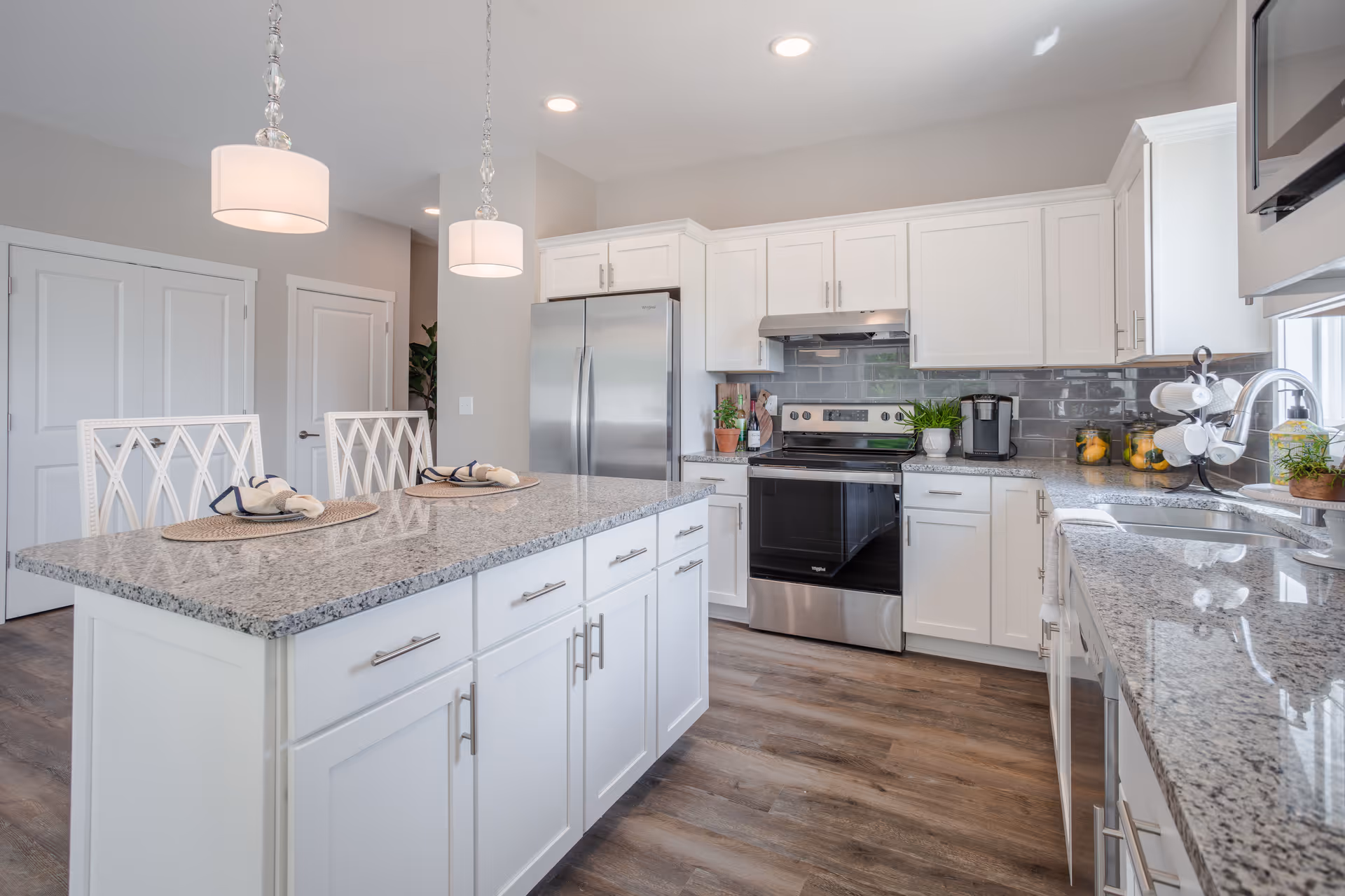 Bright modern kitchen with white cabinets, granite countertops, a stainless steel refrigerator, stove, and microwave. There is a kitchen island with two white chairs and place settings, pendant lights hanging above, and a window letting in natural light.