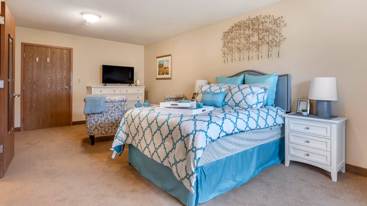 A bedroom in a senior living facility with a bed dressed in blue and white patterned bedding, a white nightstand with a lamp and small plant, a decorative metal wall art above the bed, a patterned armchair with a blue throw blanket, a white dresser with a flat-screen TV on top, and a closed wooden door.