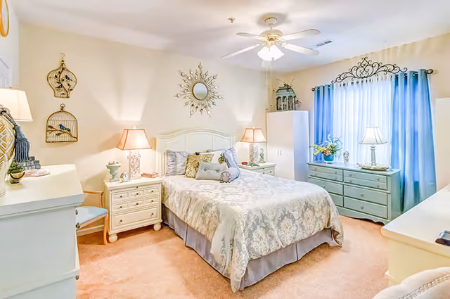 A cozy bedroom with a double bed covered in a patterned beige and white bedspread, flanked by two nightstands each with a lamp. The room features light-colored walls, a ceiling fan with a light, a dresser with a lamp and flowers near a window with blue curtains, and decorative wall hangings including a sunburst mirror above the bed.