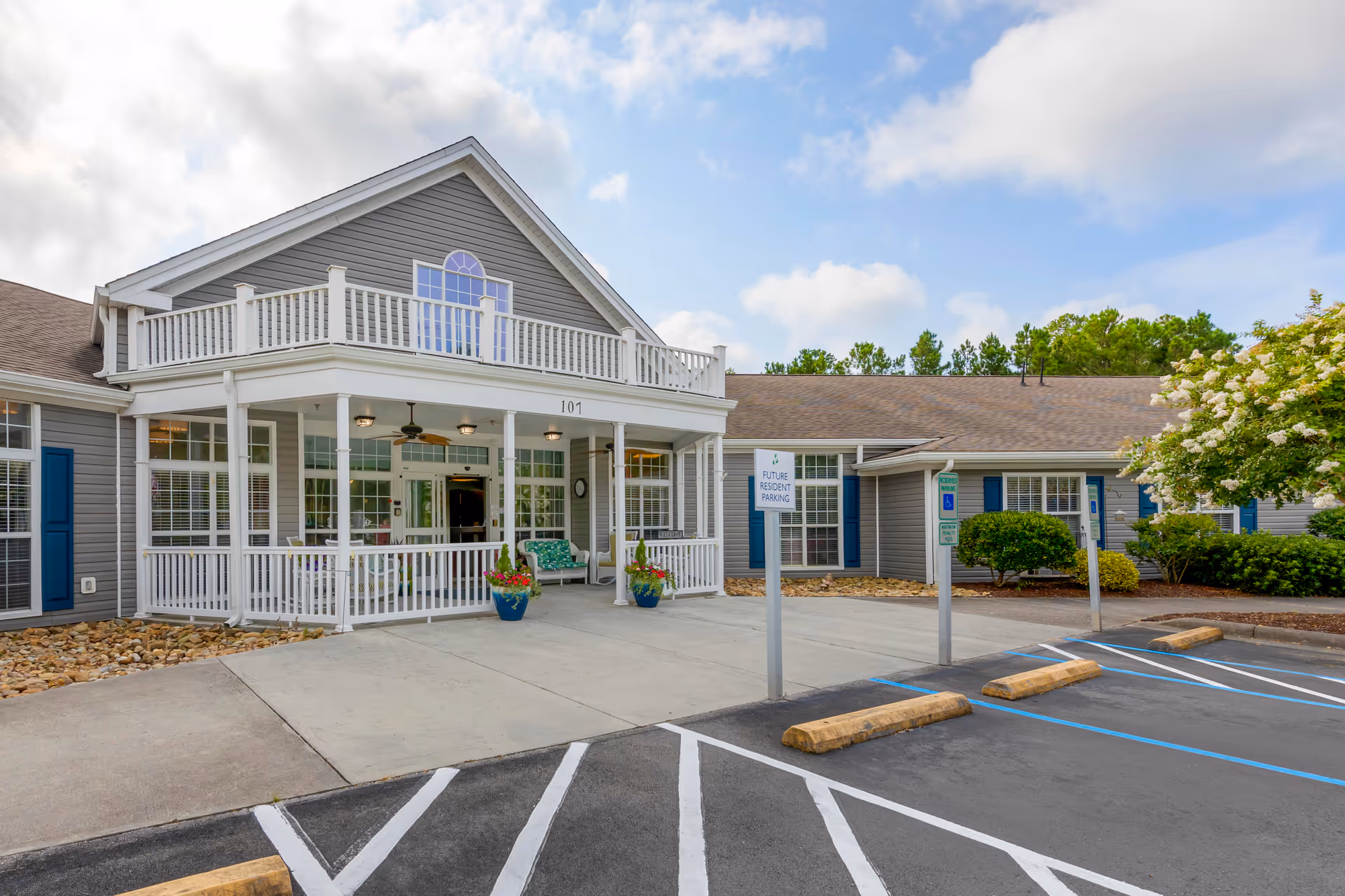 Exterior view of a senior living facility building with gray siding and white trim. The building has a covered porch with white railings, ceiling fans, and potted plants. There are parking spaces in front, including designated future resident parking spots. The sky is partly cloudy with some blue visible.