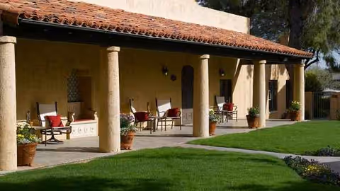 Exterior of a senior living facility with a covered patio, columns, chairs, and greenery.