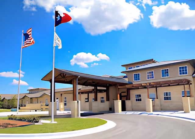 Exterior view of a senior living facility building with a covered entrance driveway. Three flagpoles are visible, flying the American flag, the Texas state flag, and another flag. The sky is blue with scattered clouds.