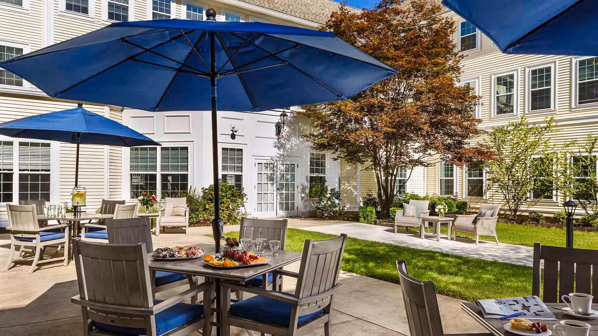 Outdoor patio area at Atria Merrimack Place featuring tables with blue umbrellas, chairs with blue cushions, a tray of food on one table, and a grassy area with additional seating and trees in front of a multi-story building with many windows.