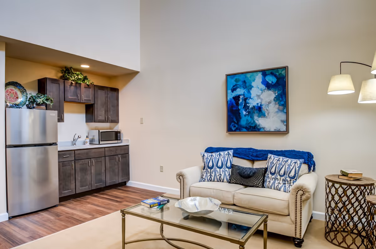 A cozy living area with a beige sofa adorned with patterned pillows and a blue throw blanket. Above the sofa hangs a blue and white abstract painting. To the right, a floor lamp with three shades provides light. In front of the sofa is a glass coffee table with a decorative bowl and a book. To the left, there is a small kitchenette with dark wood cabinets, a stainless steel refrigerator, a microwave, and a sink. The floor is a combination of wood and carpet.