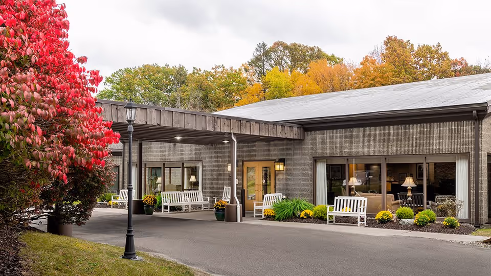 Exterior view of Elderwood Village at Vestal showing the entrance area with a covered driveway, white benches, potted plants, and a street lamp. Trees with autumn foliage in red, yellow, and green surround the building.