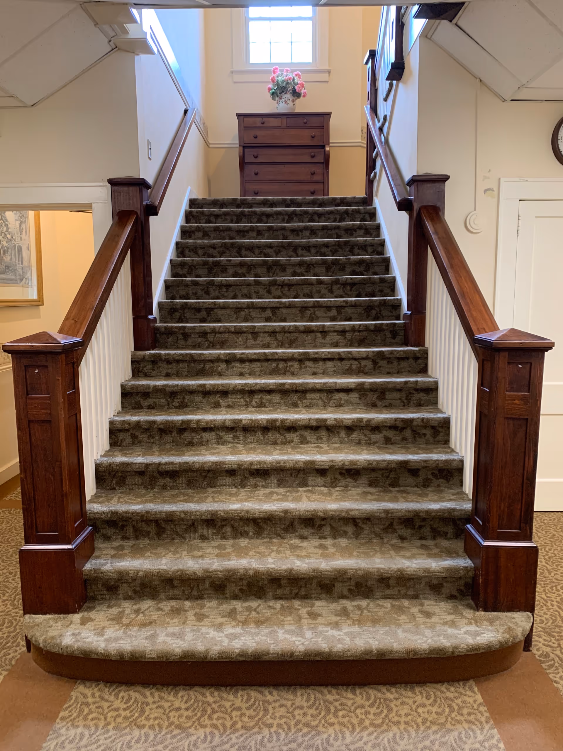 Carpeted staircase with wooden handrails on both sides leading up to a landing with a wooden chest of drawers topped with a vase of pink flowers, illuminated by a window above.