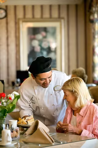 A chef wearing a white uniform and black hat is smiling and interacting warmly with an elderly woman seated at a dining table. The table has a plate of sandwiches, a napkin, and some flowers in a vase. The setting appears to be a cozy dining area with patterned wallpaper and framed artwork in the background.