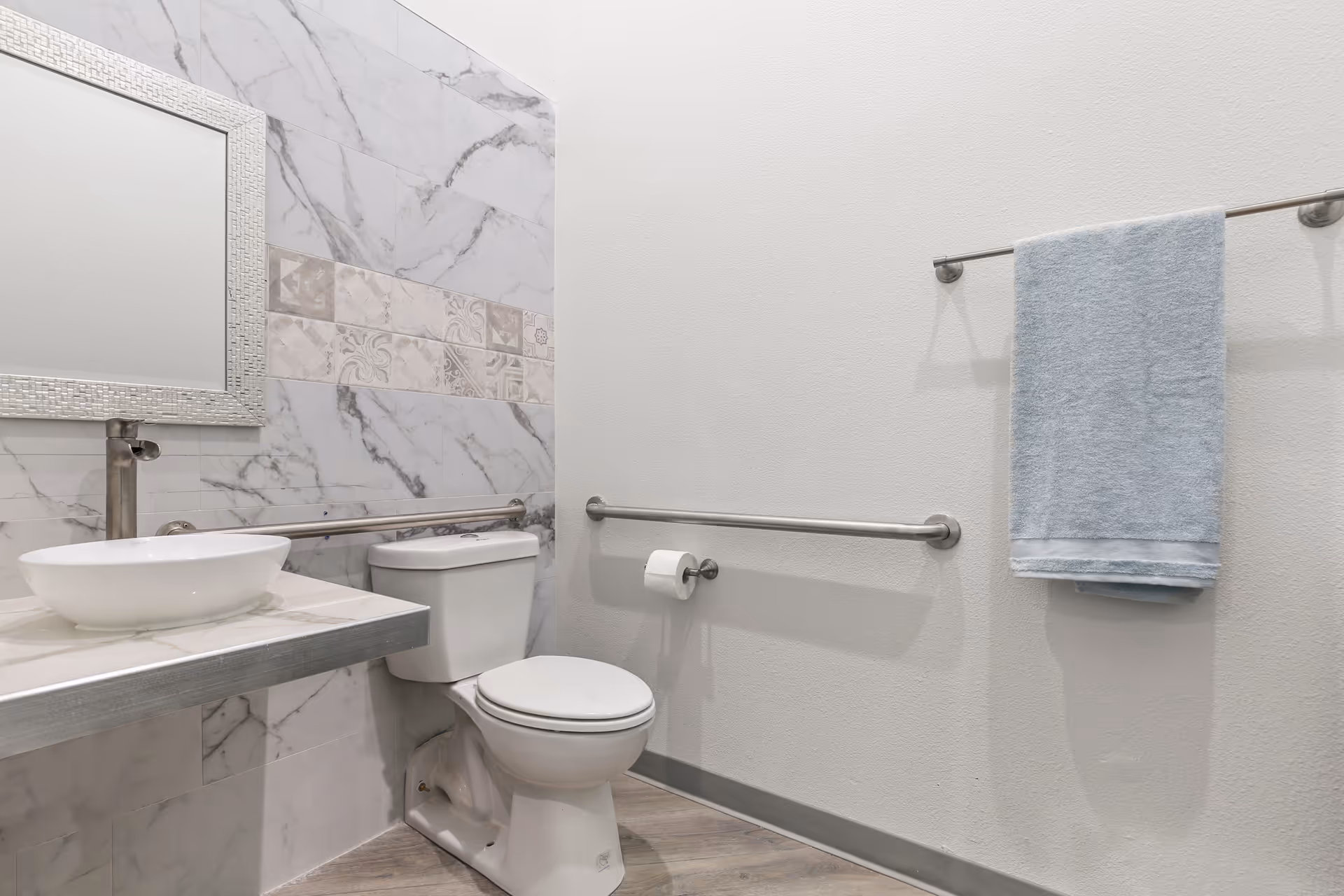 A clean and modern bathroom with a white toilet, a floating vanity with a round vessel sink, a silver faucet, and a large mirror with a textured silver frame. The walls have a marble tile design with decorative accent tiles, and there are stainless steel grab bars installed near the toilet and along the wall. A light blue towel hangs on a towel rack on the right wall.