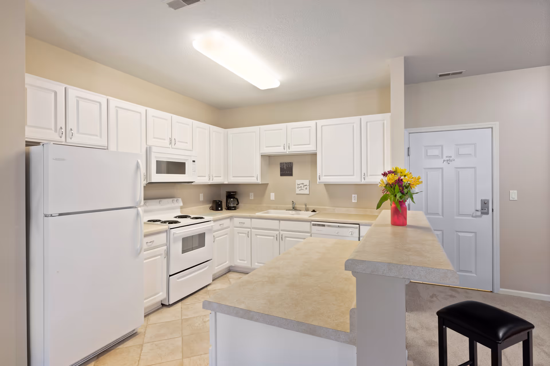 A clean, well-lit kitchen with white cabinets, a white refrigerator, stove, microwave, and dishwasher. The countertops are beige, and there is a small vase with colorful flowers on the counter. The floor is tiled, and a black stool is positioned near the counter. A door with the words 'Stay positive' is visible in the background.