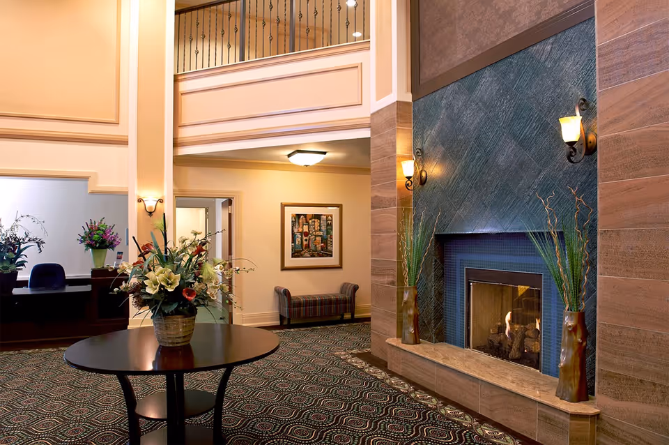 A spacious and warmly lit interior lobby area with a large fireplace on the right wall, decorated with two tall vases containing plants. In the center, there is a round wooden table with a floral arrangement. The floor is covered with a patterned carpet, and the walls are painted in soft beige tones. In the background, there is a reception desk with flowers and a seating area with a framed picture on the wall.