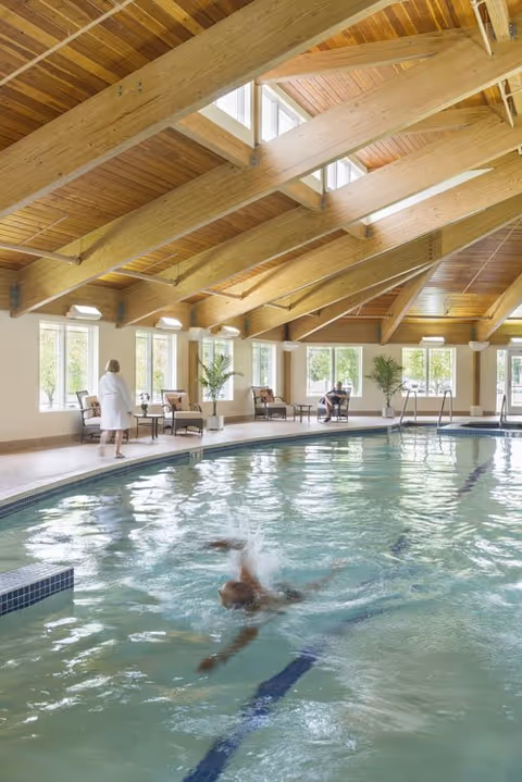 Indoor swimming pool with a wooden beam ceiling and large windows letting in natural light. A person is swimming in the pool while another person in a white robe walks along the poolside. There are chairs and plants arranged near the windows.
