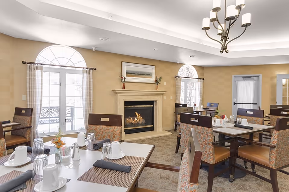 A cozy dining room with several tables and chairs arranged neatly. Each table is set with white cups, glasses, black napkins, and small flower vases. The room features a lit fireplace with a framed picture above it, two large arched windows with curtains, beige walls, and a chandelier hanging from the ceiling.