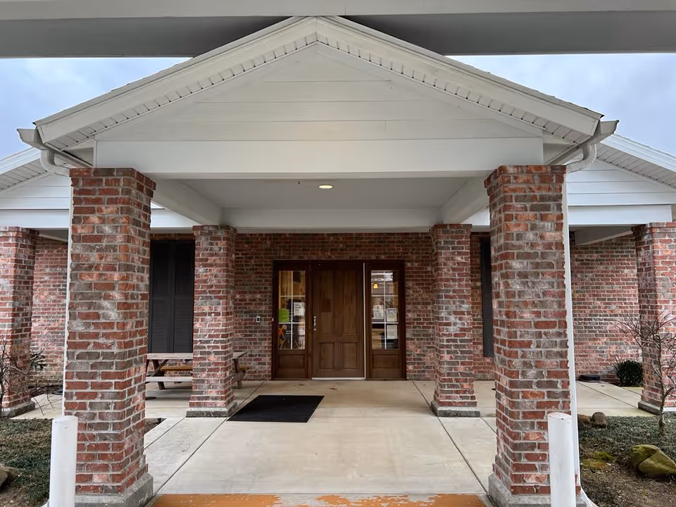 Entrance of a brick building with a covered porch supported by brick columns. The entrance has double wooden doors with glass panels on either side. There is a black doormat in front of the doors and some benches visible under the porch.