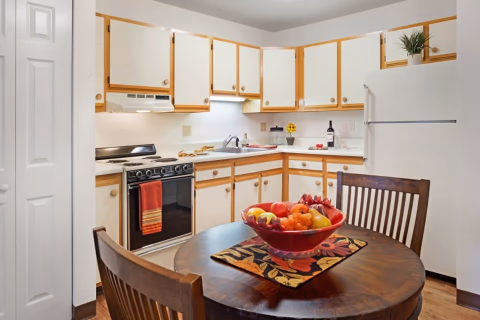 A kitchen with white cabinets and wooden trim, a white refrigerator, a stove with a red towel hanging on the handle, and a round wooden table with two chairs. On the table is a red bowl filled with various fruits and a colorful placemat underneath. There is a small plant on top of the refrigerator and some kitchen items on the counter.