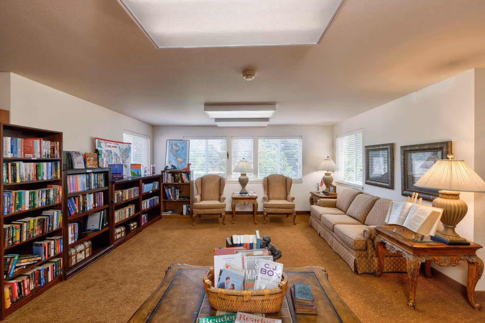 A cozy reading room with multiple bookshelves filled with books along the left wall, two beige armchairs with a small wooden table between them, a beige sofa on the right side with a wooden side table and lamp, and a coffee table in the foreground holding a basket of books. The room has large windows with blinds letting in natural light and framed pictures on the walls.