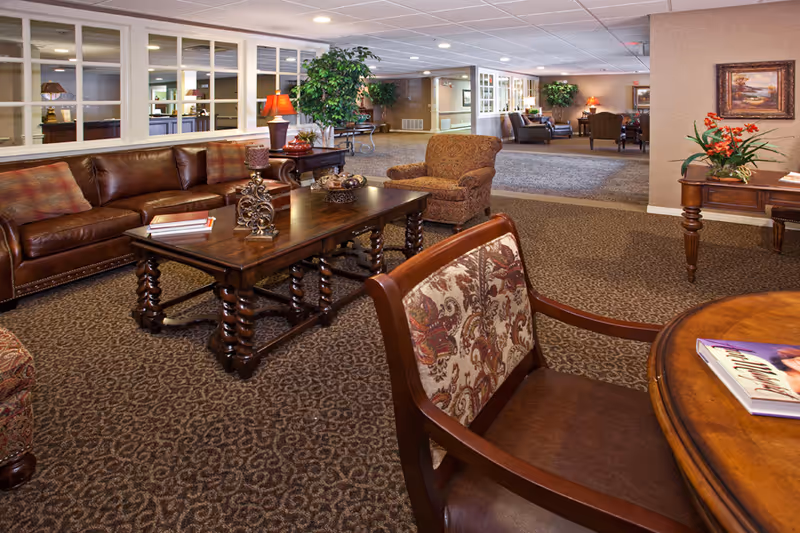 A spacious and warmly decorated assisted living common area with patterned carpet, a brown leather sofa, upholstered armchairs, wooden tables with decorative items, potted plants, and framed artwork on the walls. The room has a cozy and inviting atmosphere with soft lighting.
