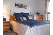 A neatly made bed with a wooden headboard and blue bedding in a bedroom. There are two wooden nightstands on either side of the bed, each with a lamp. A wooden dresser with a decorative bowl on top is against the wall near a window with sheer curtains. A framed picture hangs above the bed.