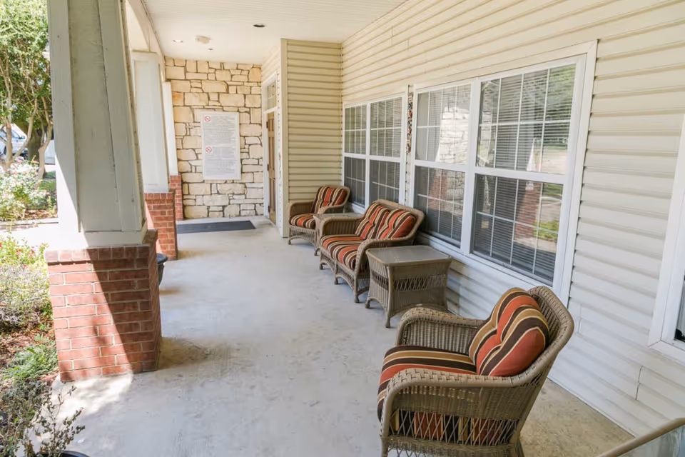 Covered outdoor porch area with four wicker chairs featuring striped cushions and a small wicker side table. The porch has a concrete floor, white siding walls, large windows with white frames, and brick columns. There is greenery and trees visible outside the porch.
