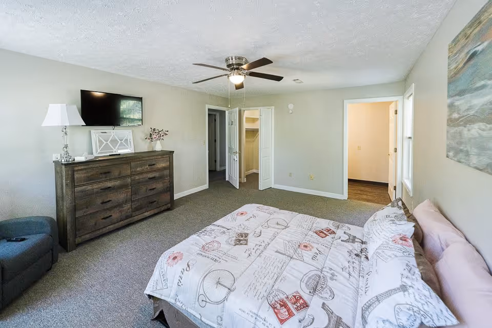 A spacious bedroom with a bed covered in a patterned quilt, a dark wooden dresser with a lamp and decorative items, a wall-mounted flat-screen TV, a ceiling fan with light, an armchair, and two open doorways leading to a closet and another room.
