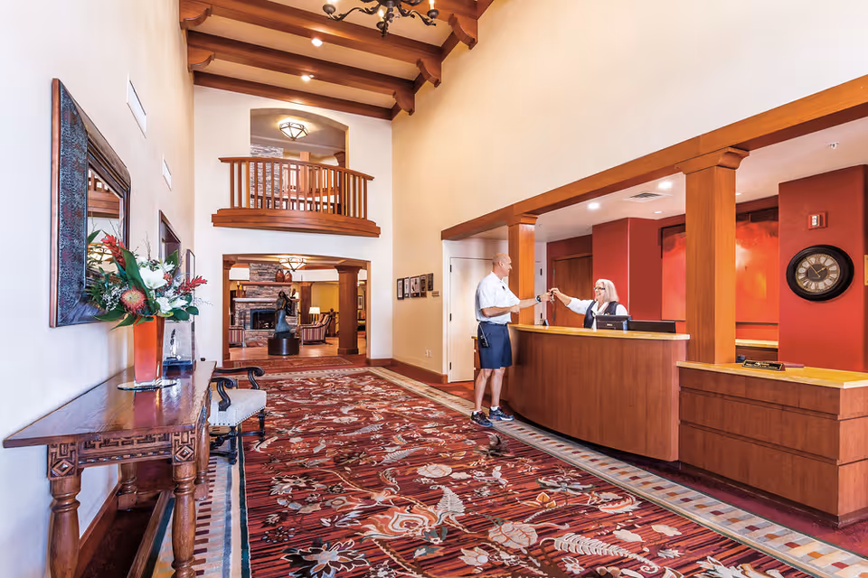 A spacious reception area in a senior living facility with a high ceiling and wooden beams. A receptionist stands behind a wooden front desk handing a key to a man in casual attire. The floor is covered with a patterned red carpet, and there is a wooden table with a floral arrangement and a large mirror on the left wall. In the background, a cozy lounge area with a fireplace and seating is visible.